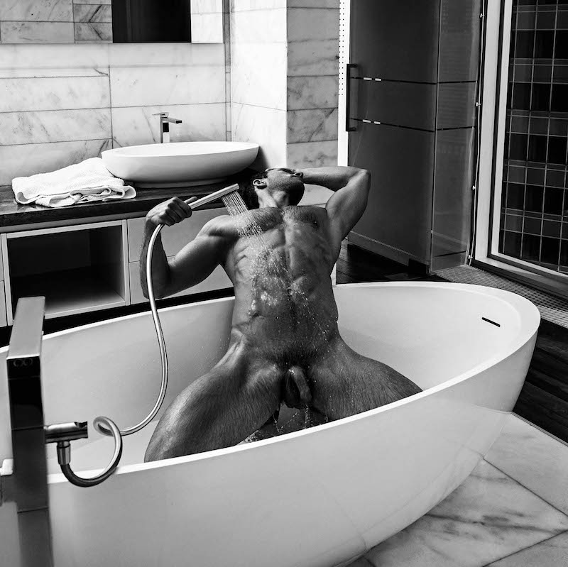 Naked male model Andrey Pavlov leaning back in a bathtub and showering himself with water while his long uncut cock hangs between his thighs.